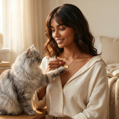 A woman in a cream linen shirt smiles while wearing a delicate gold nameplate necklace, with a fluffy gray cat resting on her shoulder and touching the necklace. The scene is set in a warm, softly lit bedroom with beige tones.