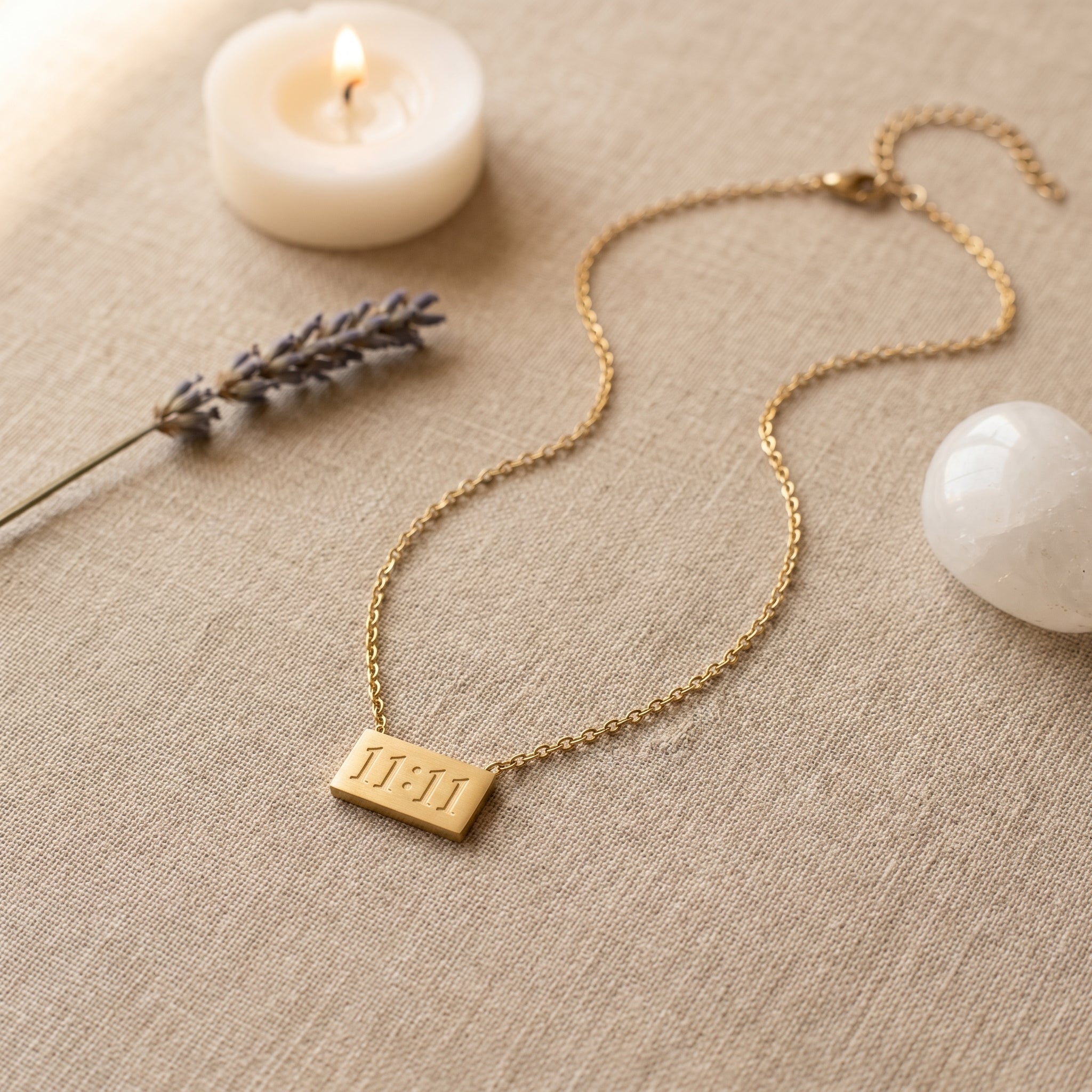 A gold chain necklace with a rectangular pendant engraved with the Roman numeral 'III' is displayed on a textured beige linen surface. The scene is styled with a lit white candle, a sprig of lavender, and a white stone.