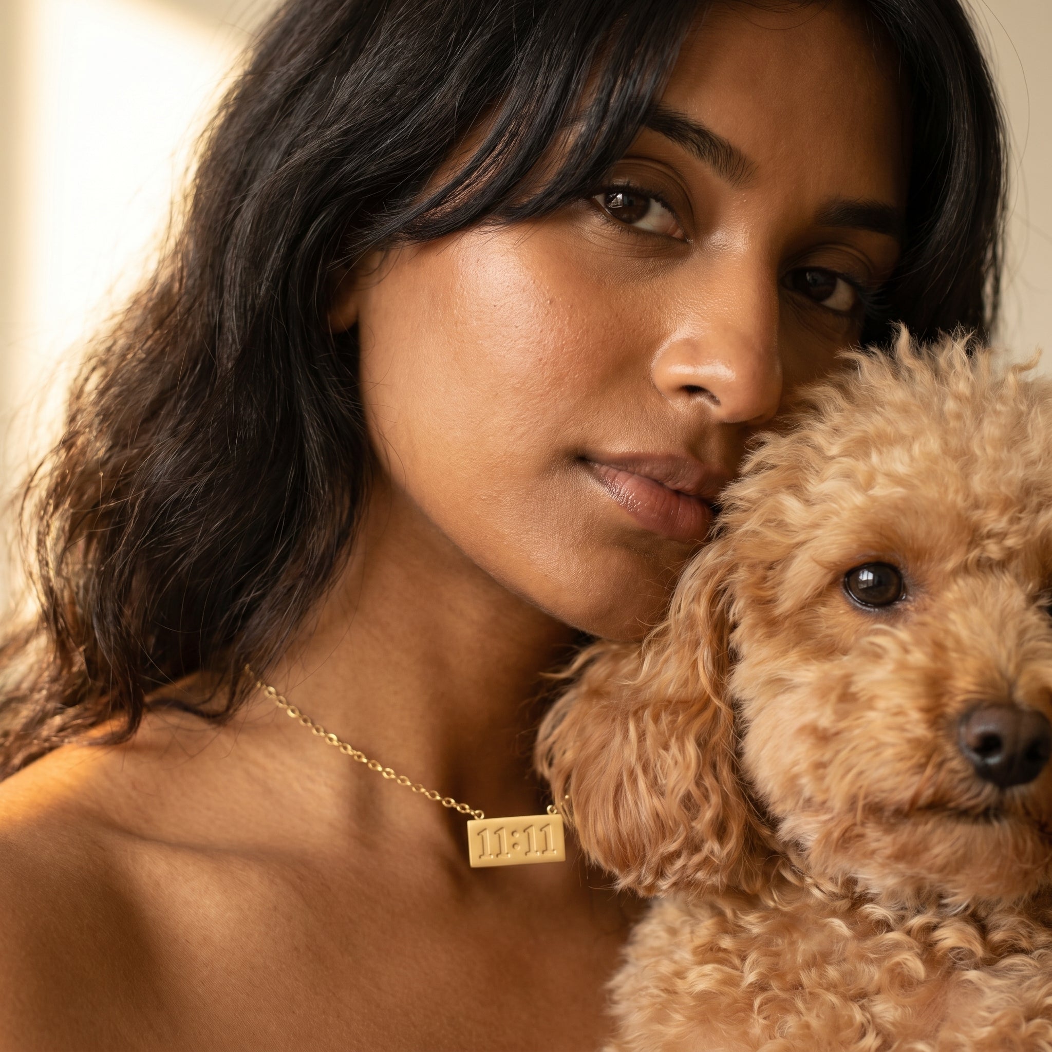 A woman with dark hair and a fluffy light brown poodle dog wearing a gold necklace with a rectangular engraved plate that reads '1311'.