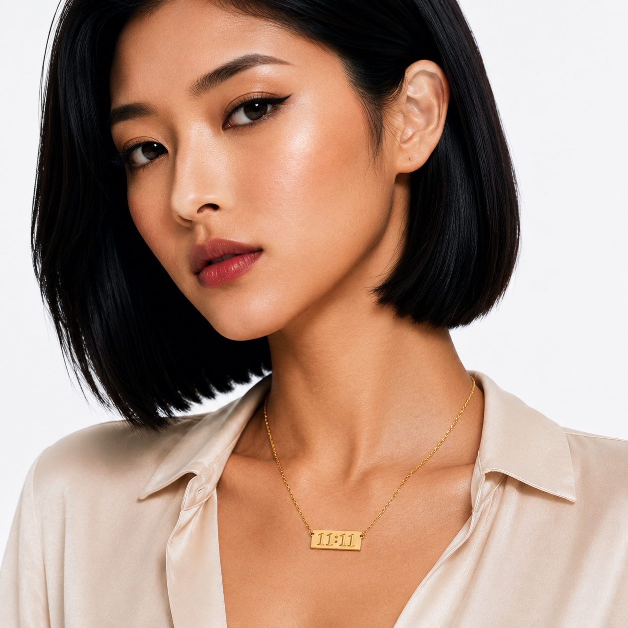 A close-up of a woman with a short dark bob hairstyle wearing a delicate gold nameplate necklace and a light beige satin shirt against a clean white background.