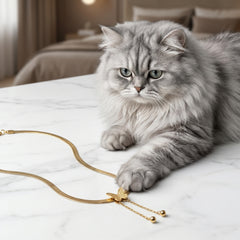 A fluffy silver-gray cat lies on a white marble tabletop, reaching toward a gold snake-chain necklace with a butterfly pendant in a softly lit bedroom setting.