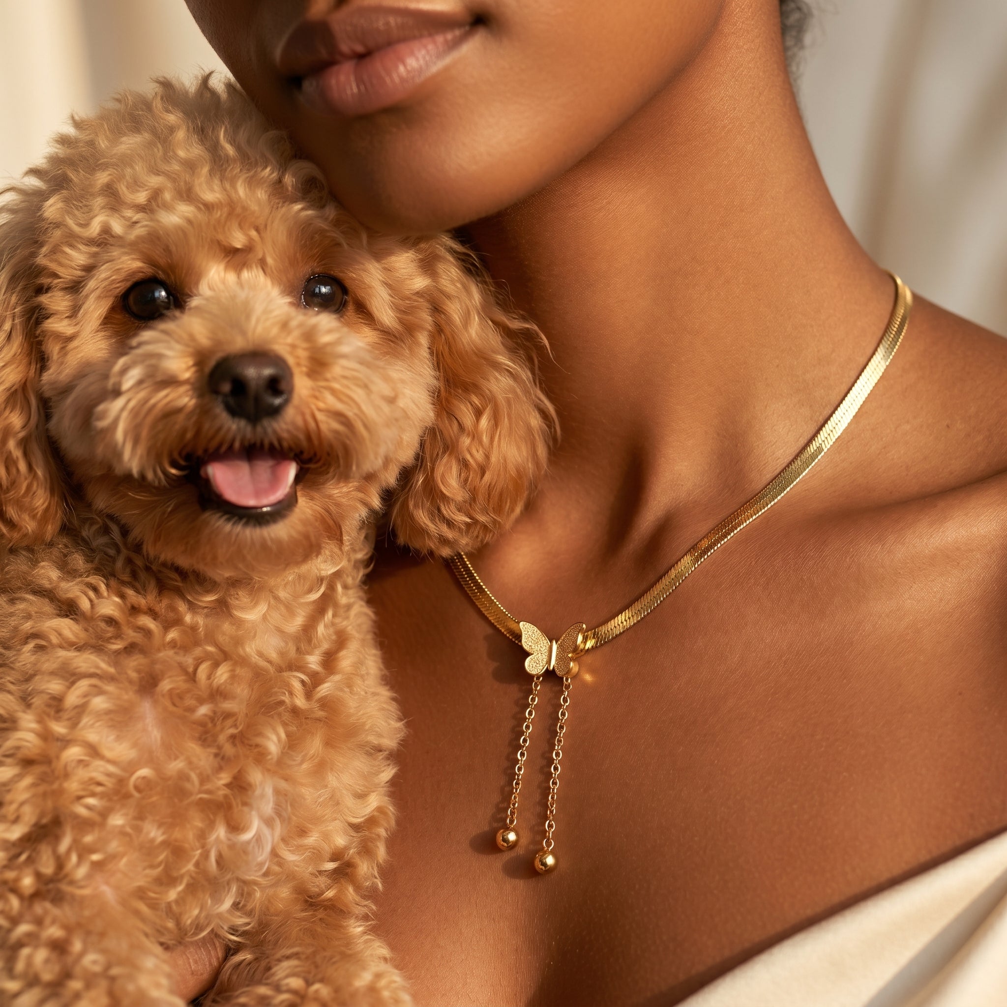 A close-up lifestyle shot of a woman wearing a gold snake chain necklace with a butterfly charm and dangling chains, while holding a small curly-haired brown dog.