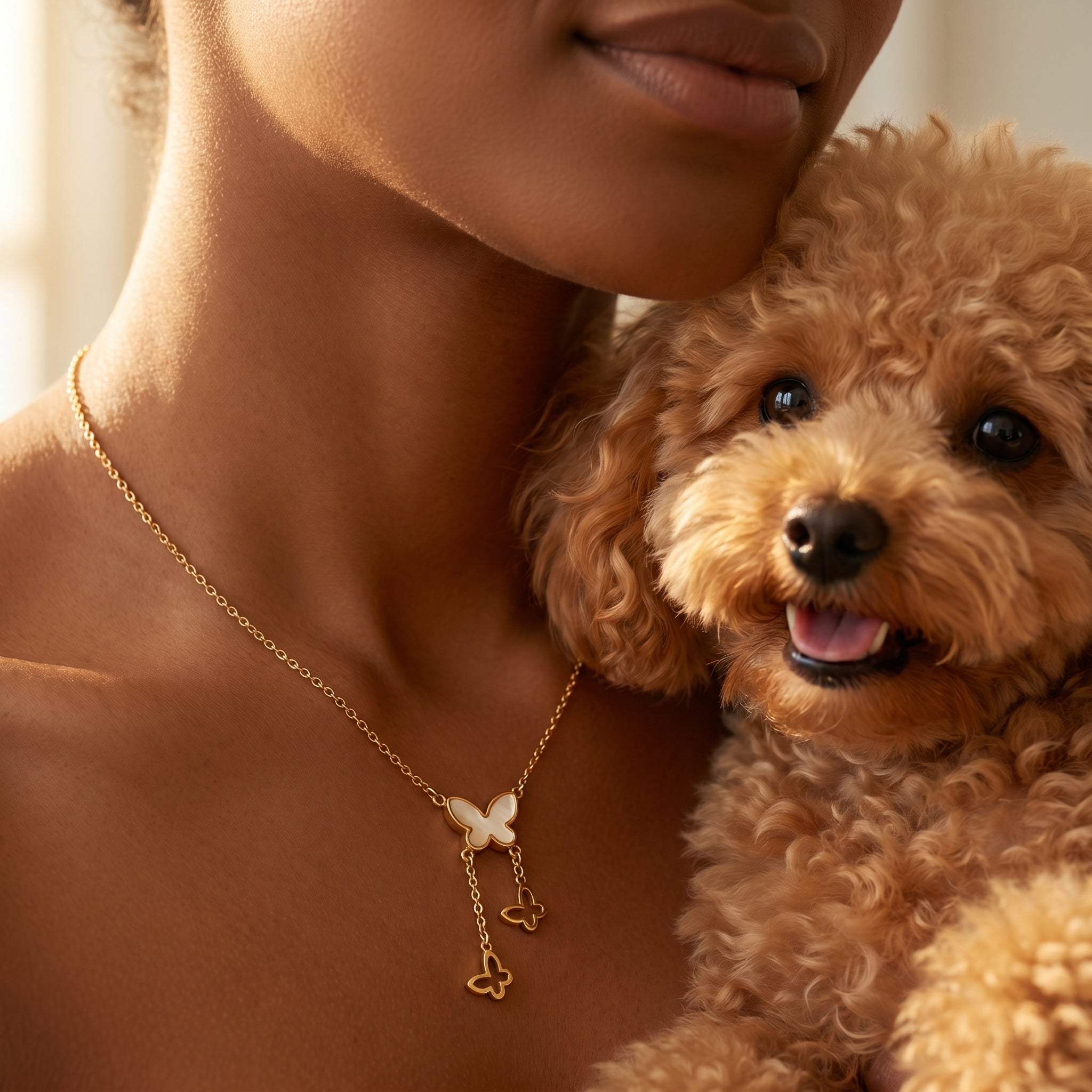 A close-up lifestyle shot of a woman holding a small curly-haired brown dog, both wearing a delicate gold necklace with a butterfly and key pendant.