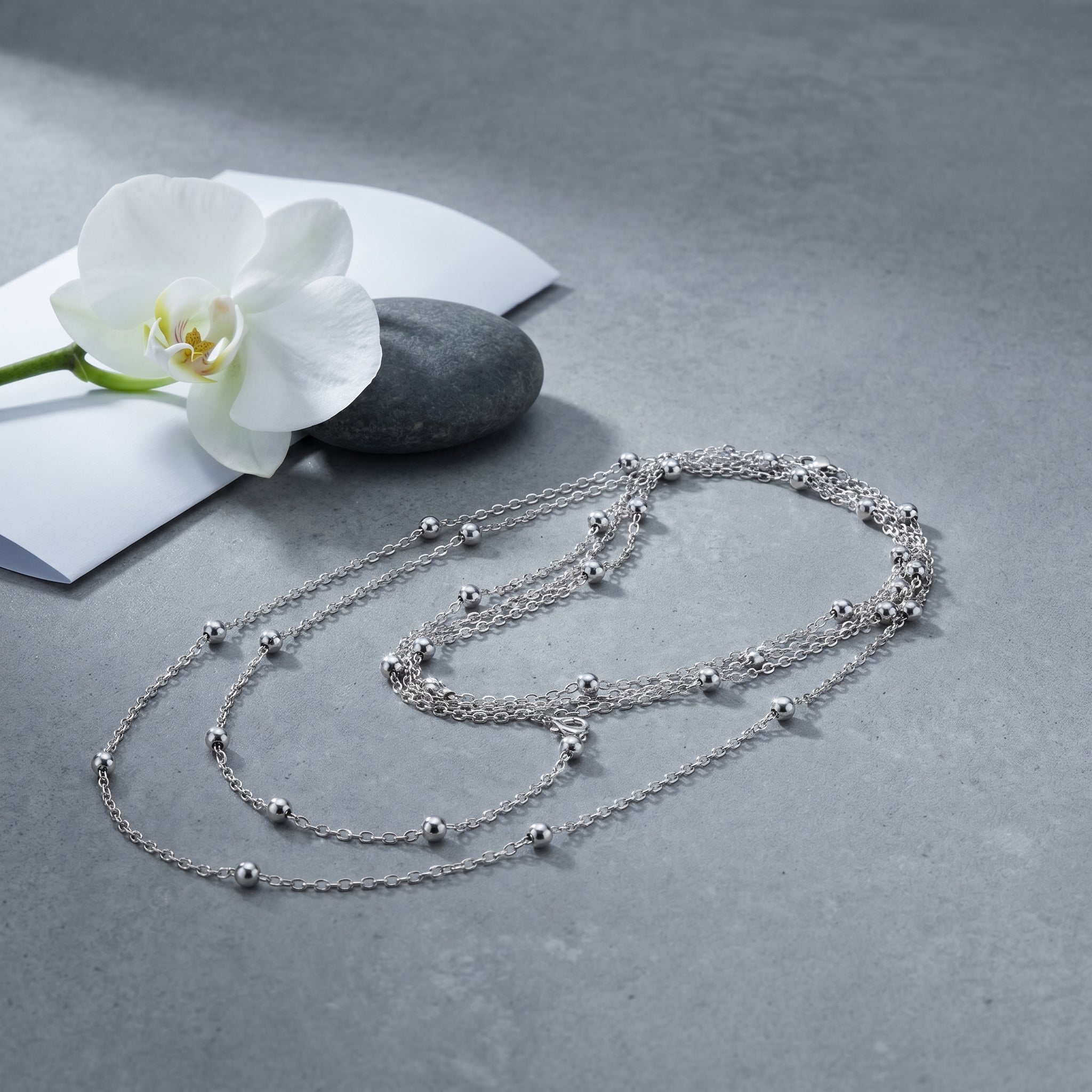 A delicate silver multi-strand necklace with small bead accents is arranged on a textured gray surface next to a white orchid flower and a dark stone.