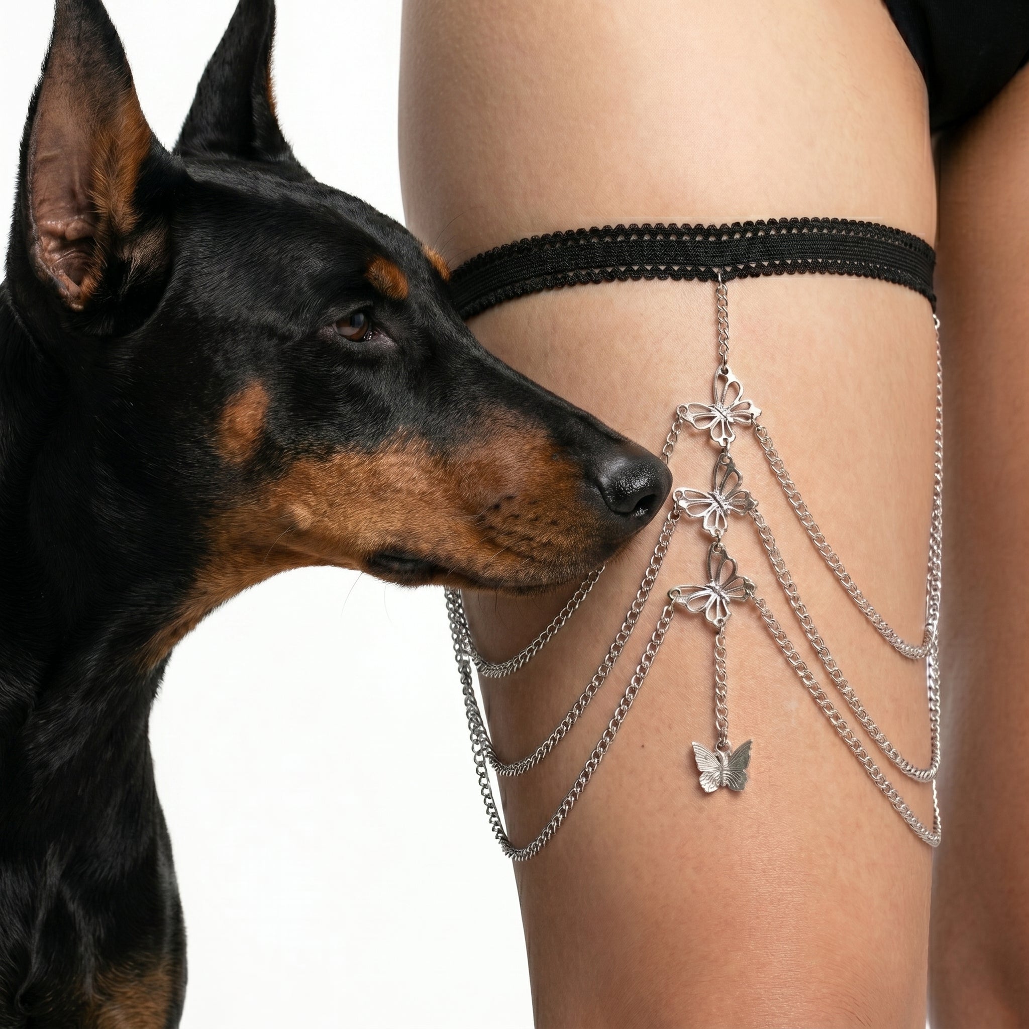 A black lace thigh strap with dangling silver chains and butterfly charms worn on a person's thigh, with a black-and-tan dog looking at the product against a white background.