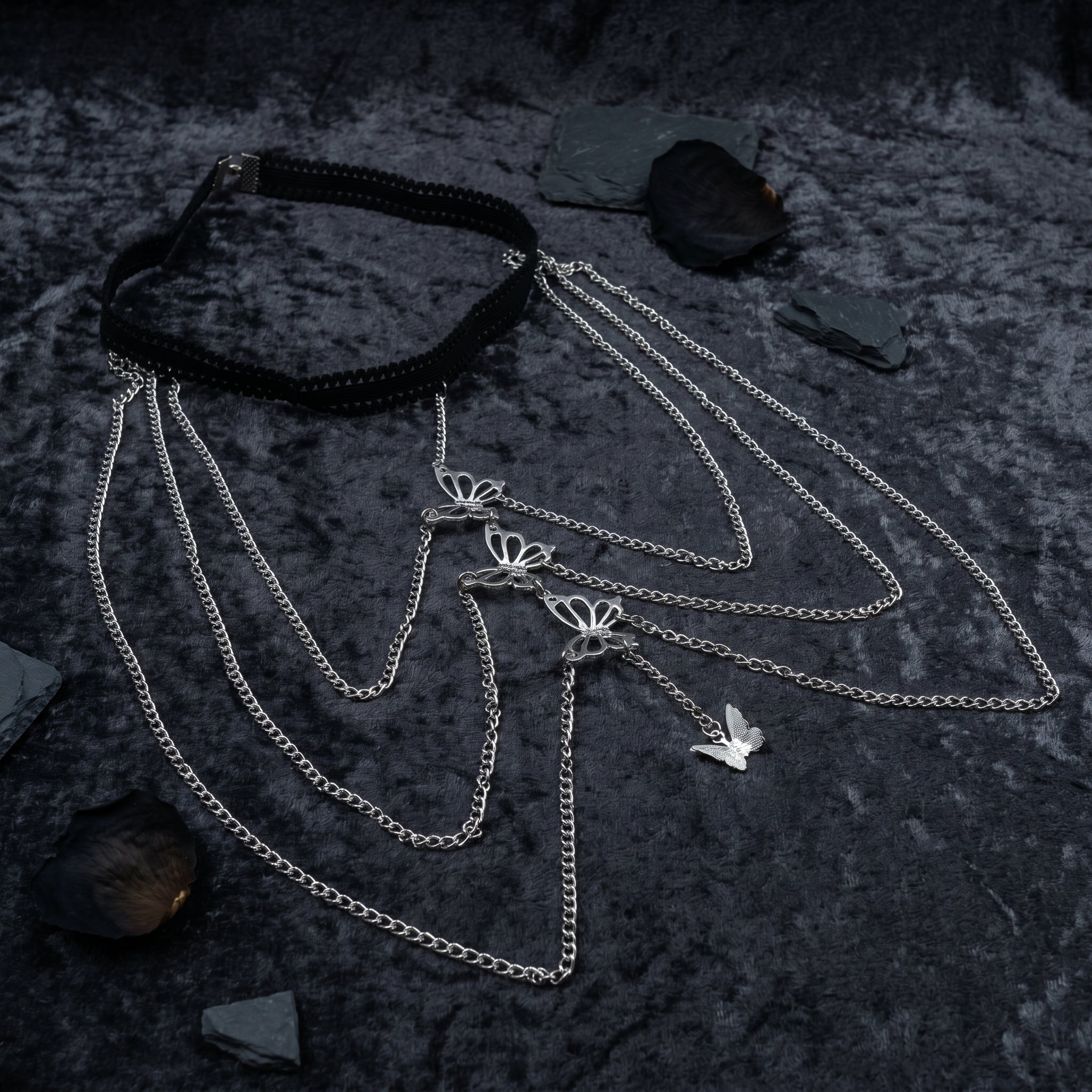 A gothic-style layered necklace featuring a black braided choker collar and multiple silver-tone chains with butterfly and bird charms, displayed on a dark textured background with scattered slate chips.