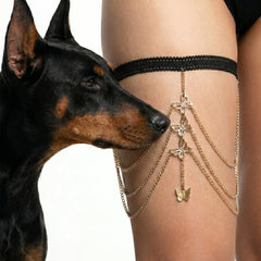 A black lace and gold chain leg strap with butterfly and flower charms worn on a person's thigh, with a black-and-tan dog looking at it from the side against a white background.