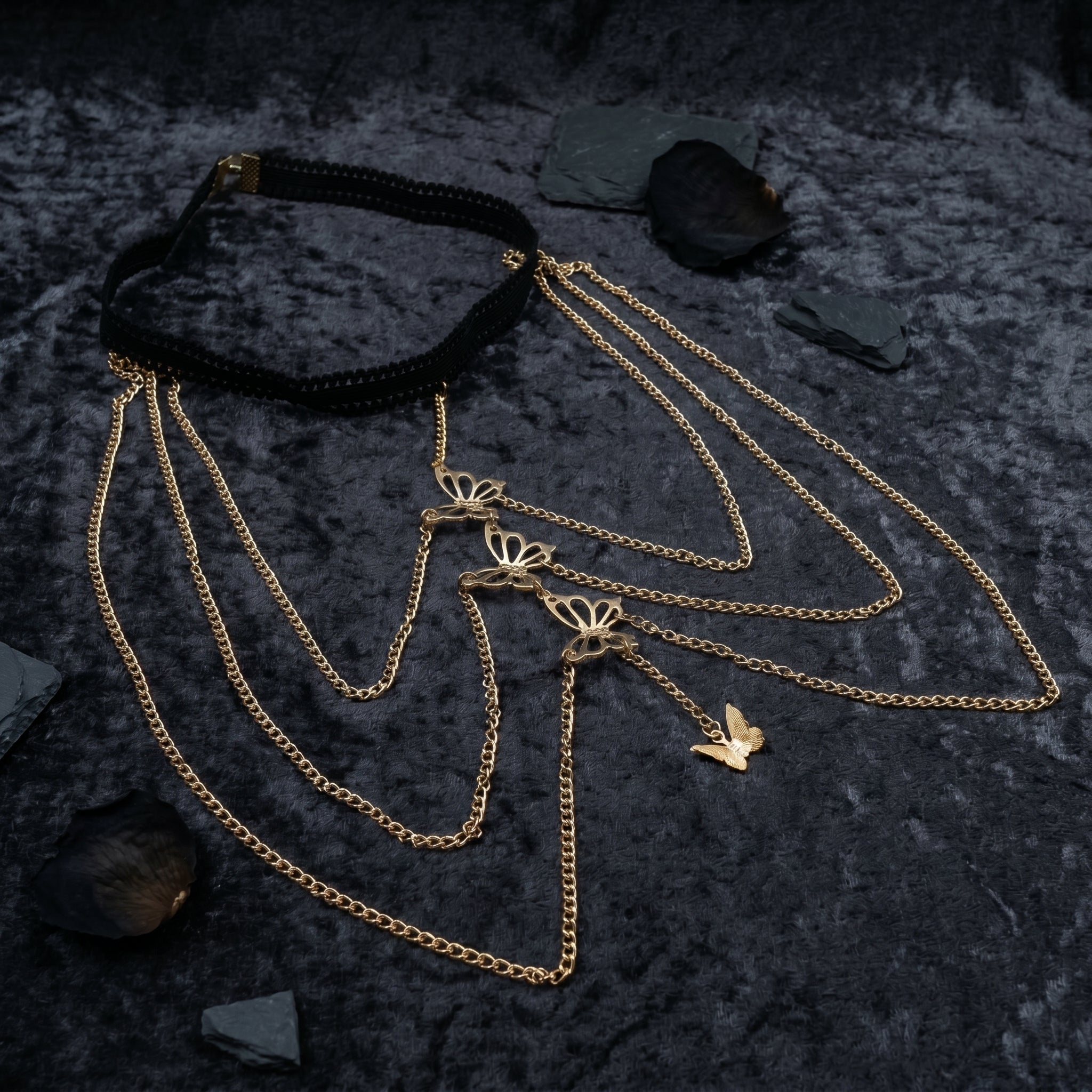 A layered gothic-style necklace featuring a black braided choker and multiple gold-tone chains with butterfly and bird charms, displayed on a dark textured background with small slate stones.