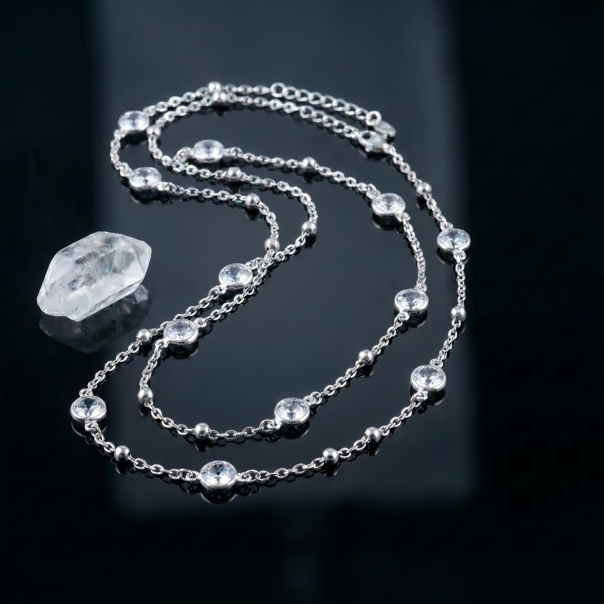 A silver-toned chain necklace with multiple clear faceted gemstones and a larger clear crystal pendant, displayed on a glossy black reflective surface.