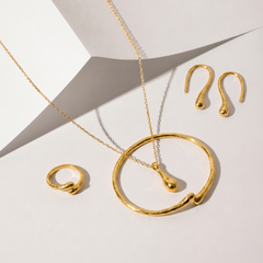 Gold jewelry set featuring a necklace with a large open circular pendant, a matching ring, and two matching teardrop-shaped earrings, arranged on a white geometric background.