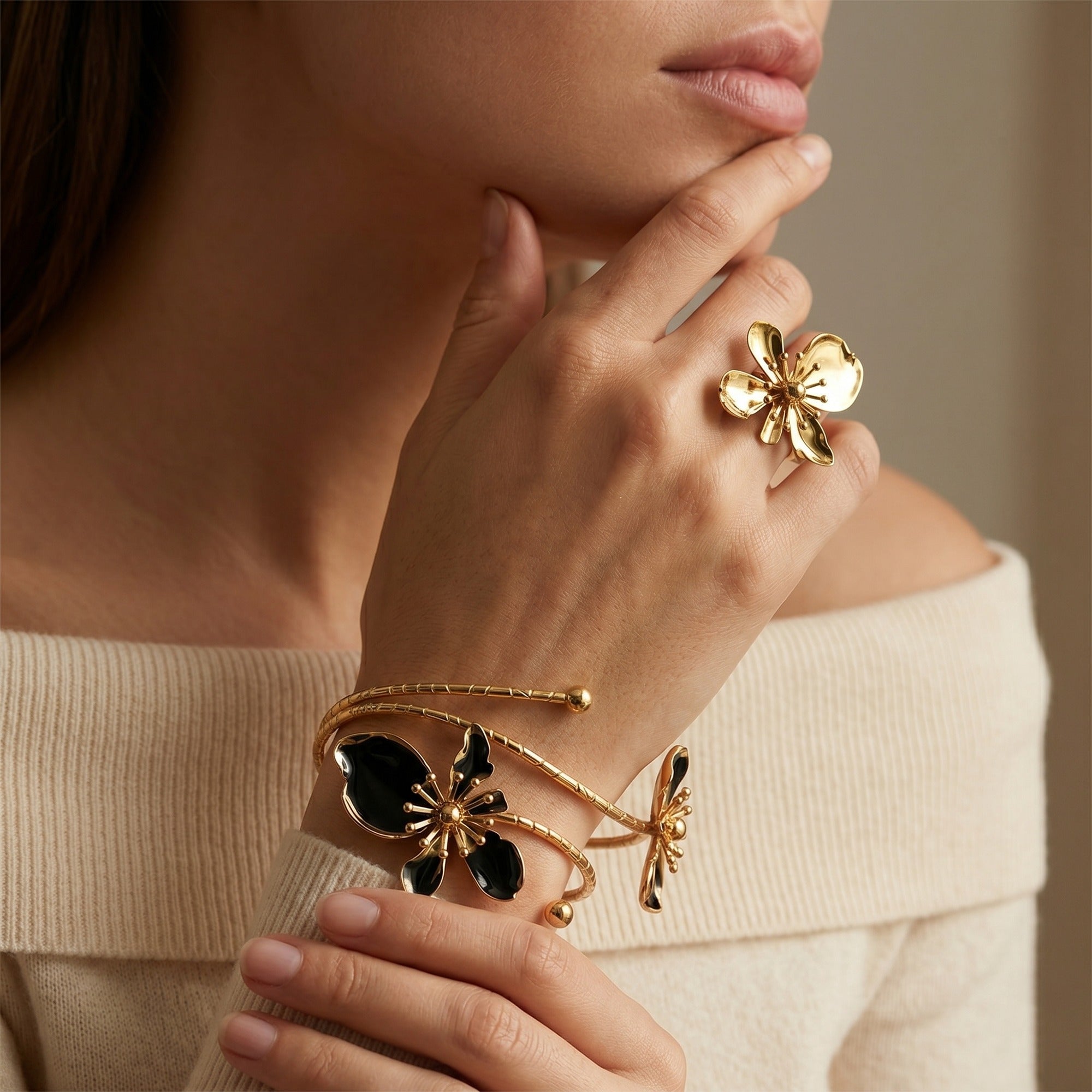 A close-up lifestyle shot of a model wearing gold jewelry, featuring a gold ring shaped like a flower and a gold cuff bracelet with black enamel butterfly and dragonfly details.