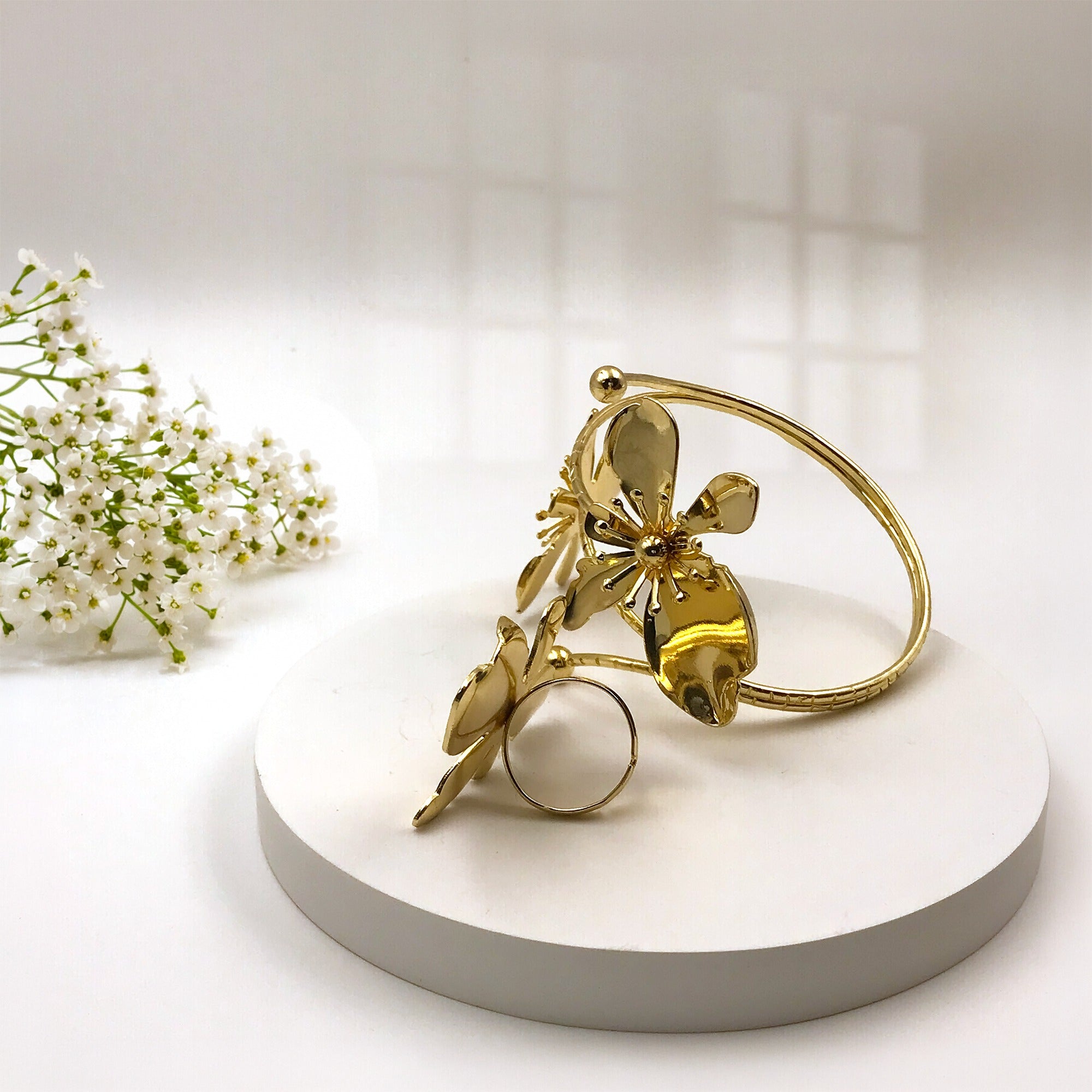 A gold-toned metal ring featuring a stylized butterfly and leaf design, displayed on a round white pedestal with small white flowers in the background.