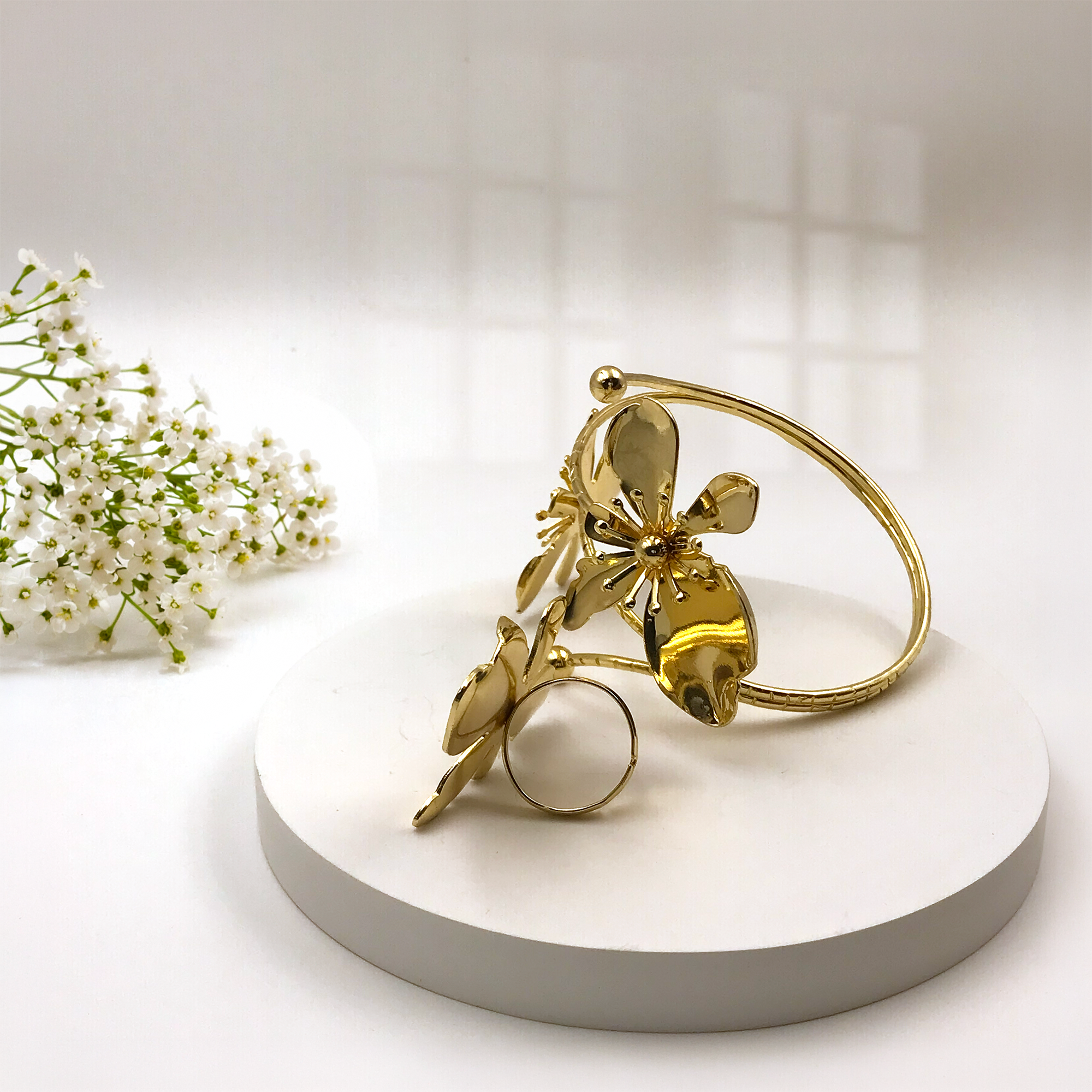 A gold-toned metal ring with a sculptural floral design, displayed on a round white pedestal with small white flowers in the background.