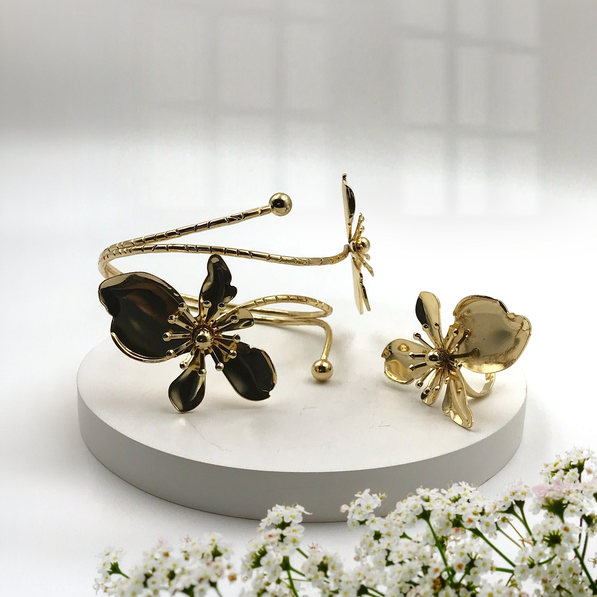 Gold-tone flower-shaped cuff bracelet and matching flower-shaped ring displayed on a round white pedestal, with small white flowers in the foreground and a softly lit neutral background.