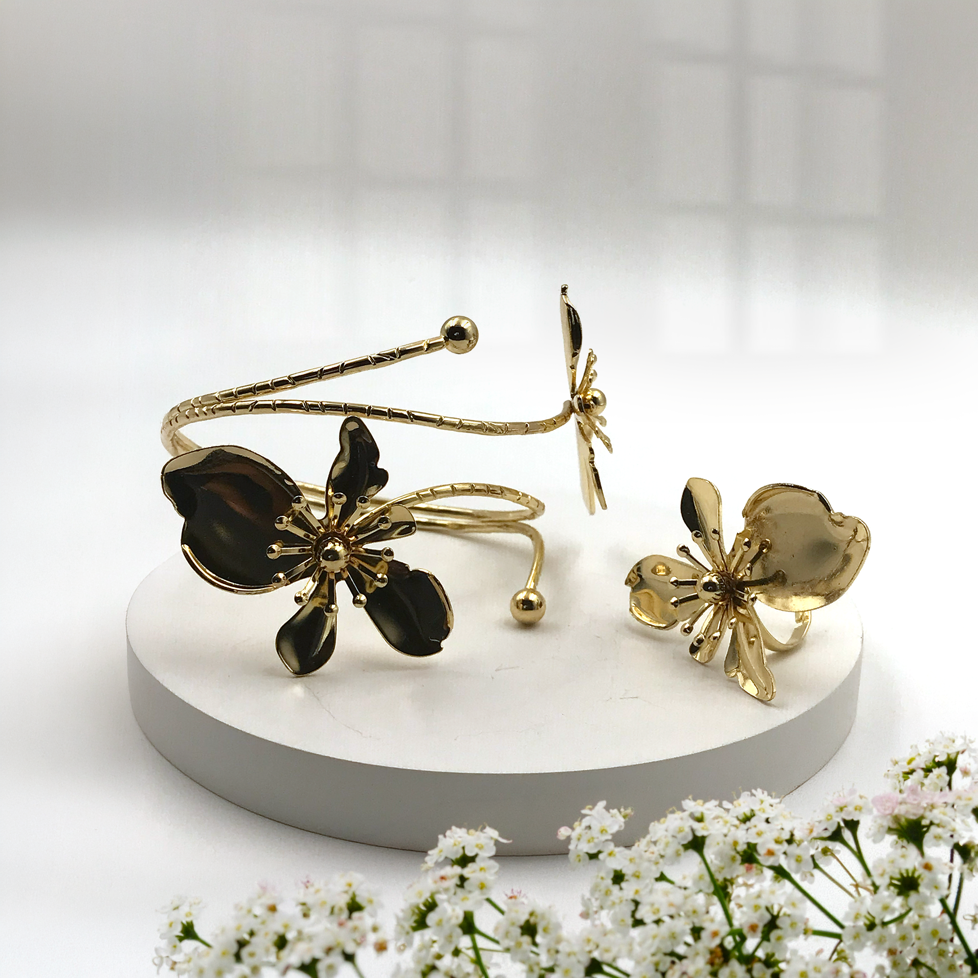Gold-tone flower-shaped cuff bracelet and matching flower-shaped ring displayed on a round white pedestal, with small white flowers in the foreground and a softly lit neutral background.