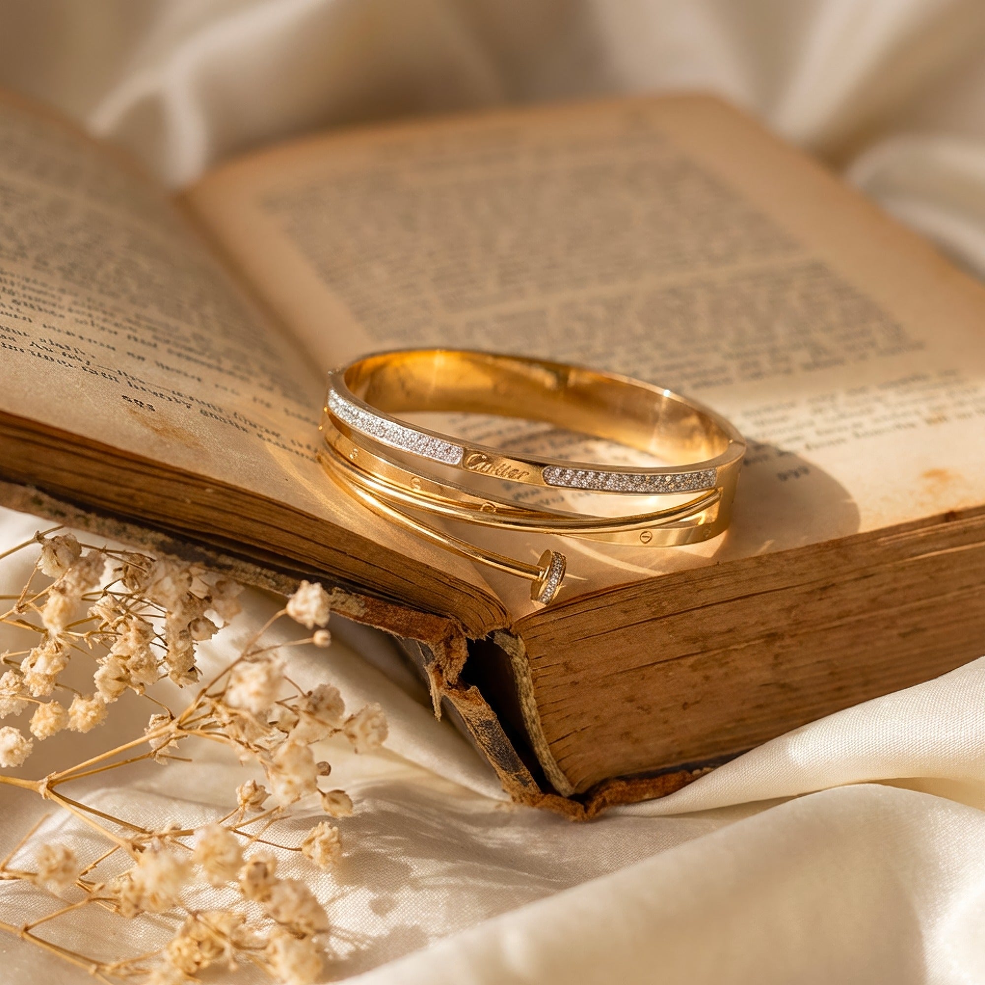 A gold bangle bracelet with a row of small clear stones across one end, resting on an open vintage book with cream-colored silk fabric and dried flowers around it.