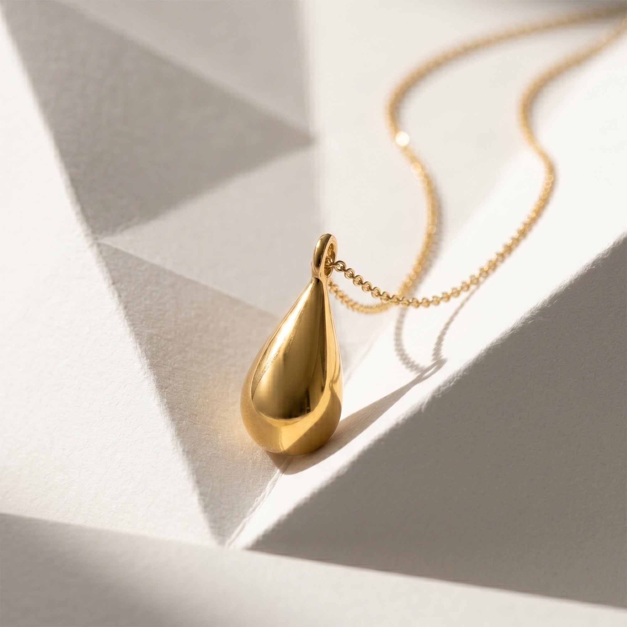 A polished gold teardrop pendant necklace on a delicate gold chain, styled on a white geometric surface with sharp shadows.