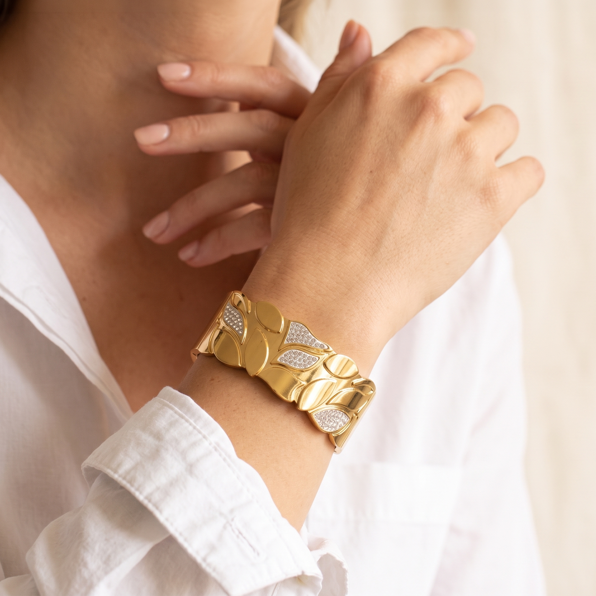 A close-up of a person wearing a wide gold cuff bracelet with leaf-like segments and pavé-set clear stones, paired with a white collared shirt.