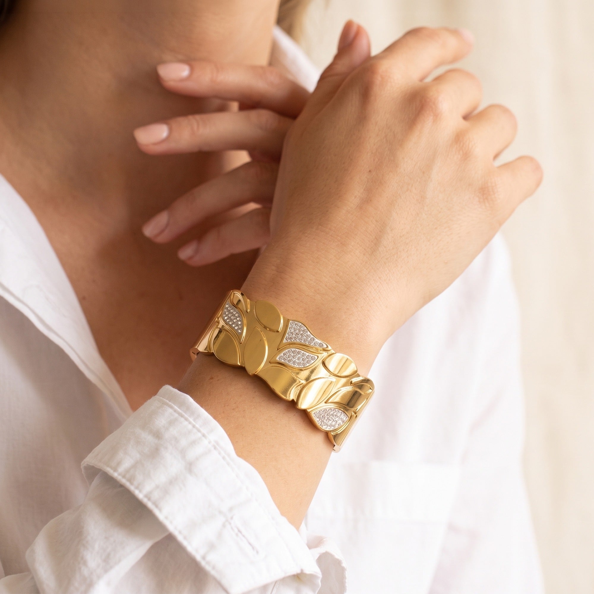 A close-up lifestyle shot of a woman wearing a wide gold cuff bracelet with textured organic shapes and pavé-set clear stones on the front, paired with a white collared shirt.