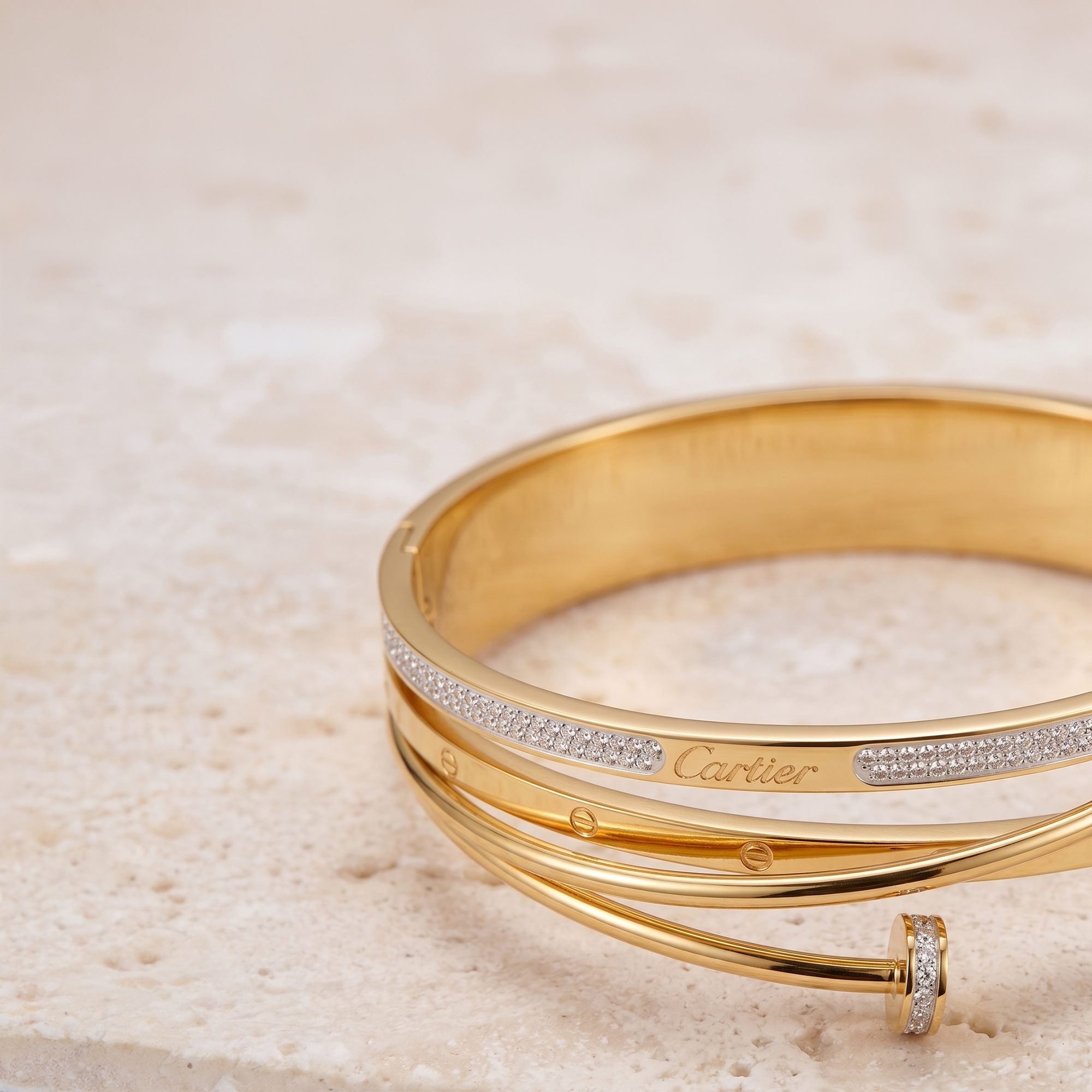 A gold bangle bracelet with a row of small clear stones and a visible Cartier logo, photographed on a light beige stone surface.