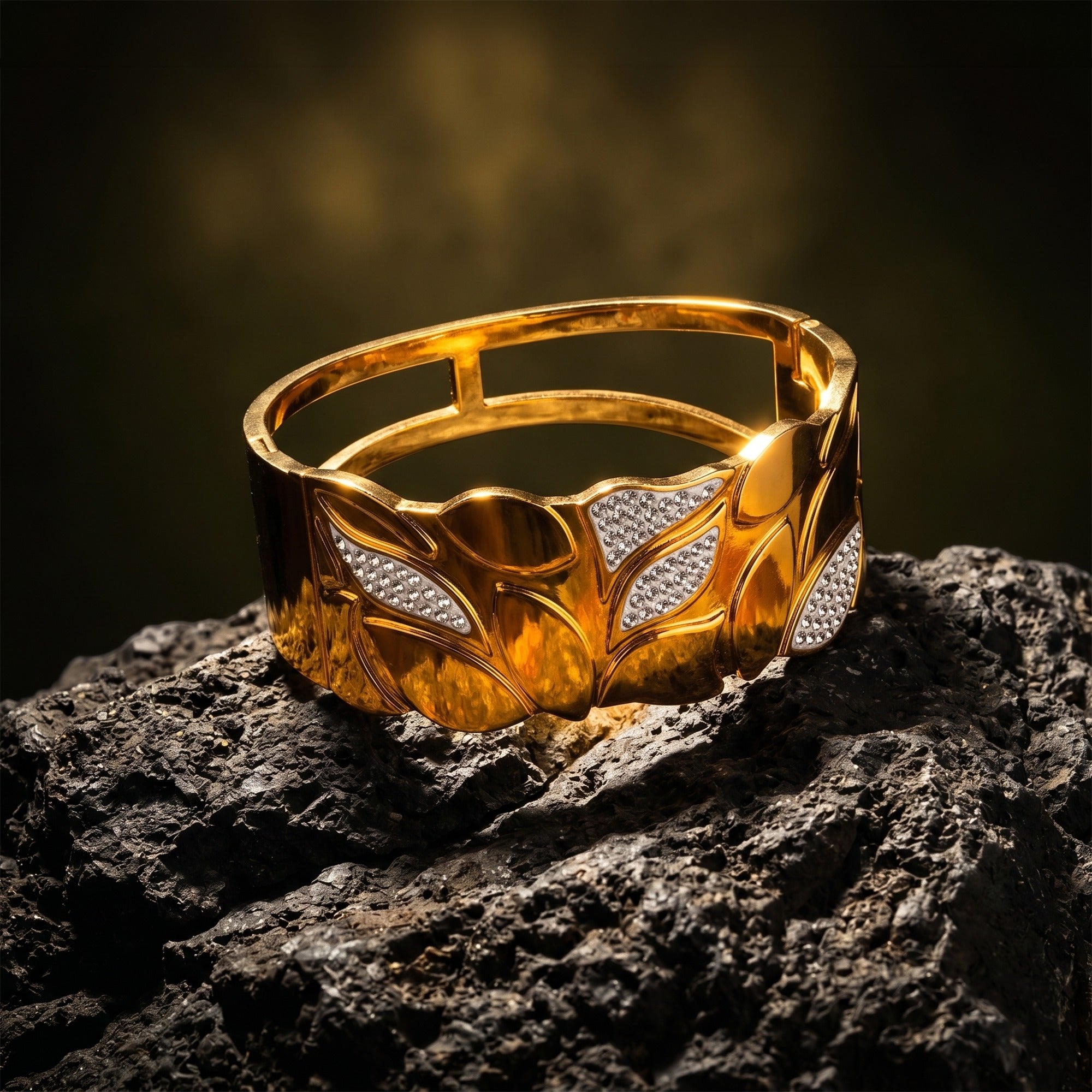A wide gold cuff bracelet with a leaf pattern and diamond-set accents, displayed on a dark textured rock against a dark, moody background.