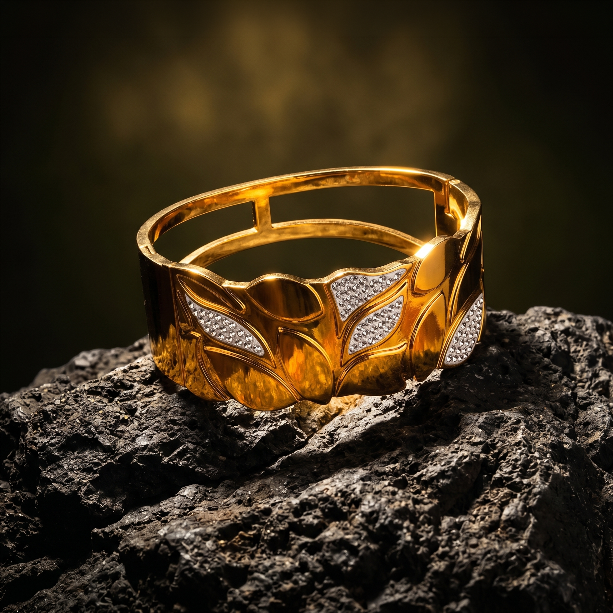 A wide gold cuff bracelet with a leaf pattern and pavé-set clear stones, displayed on a dark textured rock against a dark, moody background.